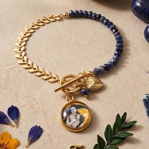 Könnte beinhalten: Ein Armband mit einer goldfarbenen Fischgrätenkette auf der einen Seite und dunkelblauen facettierten Perlen auf der anderen. Es verfügt über einen goldenen Knebelverschluss, einen kleinen Herzanhänger und einen runden Anhänger mit Frida Kahlo.