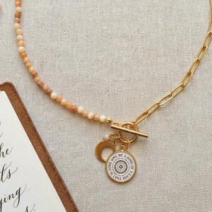 Op de afbeelding: Een ketting met een helft van kleine, ronde, gevarieerde bruine en beige kralen en een helft van goudkleurige, langwerpige ovale schakels. De ketting heeft een goudkleurige knevelsluiting met een halvemaanvormige bedel en een ronde hanger.