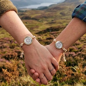 Op de afbeelding: Twee bijpassende armbanden met een ronde zilverkleurige bedel met een centraal mandala-patroon, bevestigd aan een lichtbeige gevlochten koord met kleine decoratieve kwastjes aan de uiteinden, gedragen om twee polsen die elkaar vasthouden.