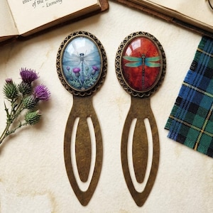 May include: Two antique bronze metal bookmarks with oval cabochons. The left bookmark features a blue dragonfly over purple thistle flowers. The right bookmark displays a dragonfly with teal wings against a deep red background. Both have open handles.