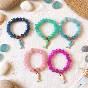 Può includere: Cinque braccialetti elastici realizzati con perline sfaccettate in blu scuro, verde brillante, azzurro, rosa pallido e magenta. Ogni braccialetto presenta un piccolo ciondolo a forma di pesce appeso con un anello in metallo color rame.
