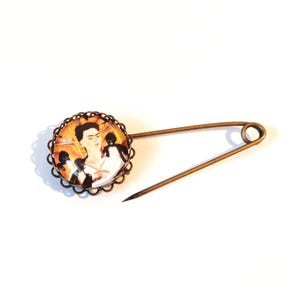Può includere: Una spilla da balia color bronzo con un'incastonatura circolare a cabochon dal bordo smerlato. Il centro mostra un ritratto colorato di una persona con capelli scuri e un gatto nero su uno sfondo dai toni caldi, protetto da una cupola di vetro.