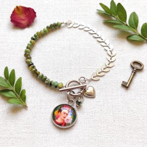 Könnte beinhalten: Ein Armband mit einer Reihe grüner, gesprenkelter Perlen und einer silberfarbenen Metallkette in Blattform. Das Schmuckstück verfügt über einen Knebelverschluss, einen kleinen herzförmigen Anhänger und einen runden Anhänger mit einem Blumenporträt.