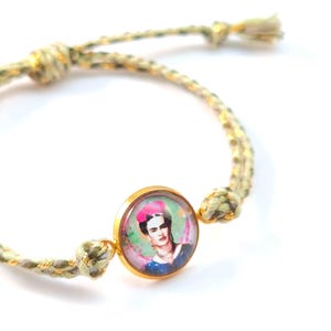 Könnte beinhalten: Ein Armband mit einem runden, goldfarbenen Metallanhänger, der ein Porträt von Frida Kahlo mit einem rosa Blumenkopfschmuck zeigt. Der Anhänger ist an einem geflochtenen Band aus hellgrünen, beigen und goldfarbenen Fäden mit verstellbaren Knoten befestigt.