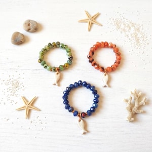 Può includere: Tre anelli di perline con pietre sfaccettate in verde, arancione e blu. Ogni anello include un piccolo ciondolo bianco a forma di pesce sospeso alla fascia. Gli anelli sono disposti in una formazione triangolare con stelle marine.
