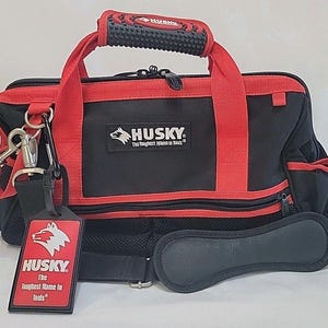 Puede incluir: Una bolsa de herramientas negra y roja con el logotipo de Husky y el texto The Toughest Name in Tools. La bolsa incluye un asa de goma texturizada, un compartimento principal con cremallera y una correa de hombro extraíble con etiqueta Husky.
