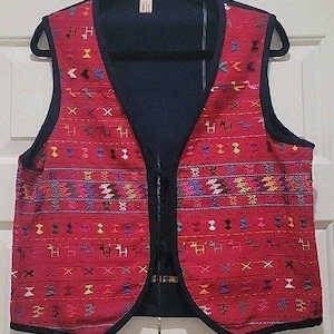 May include: A red vest with a detailed woven pattern in various colors, including blue, purple, and yellow. The vest has a black lining and trim, and a black zipper closure. The garment is displayed on a black hanger.