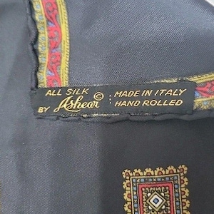 May include: A black silk scarf with a hand-rolled edge and a decorative border featuring red, blue, and gold patterns. A black fabric label is stitched onto the scarf, reading: ALL SILK BY Ashear MADE IN ITALY HAND ROLLED. A small square motif is visible.