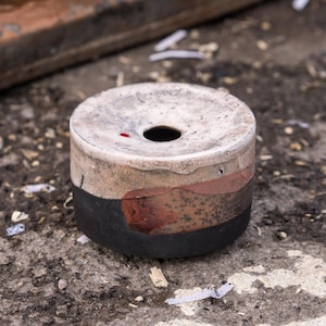 Peut inclure: Un cendrier cylindrique avec un trou central. Le cendrier a une base noire, une bande brun-rouge et un dessus de couleur claire. Il repose sur une surface grise rugueuse.