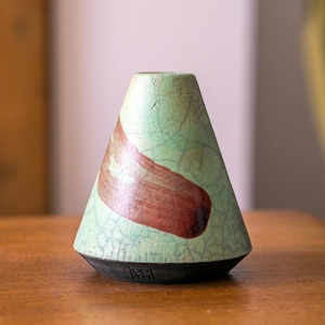 May include: A handcrafted ceramic vase with a conical shape. The vase features a light green crackle glaze with a large, reddish-brown abstract design. The base is black, and the vase is sitting on a wooden surface.
