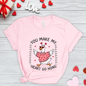 May include: A light pink t-shirt featuring a cartoon goose wearing a pink sweater with red hearts and sunglasses. The text "YOU MAKE MY HEART GO HONG" is printed around the goose in a circular design. Small red hearts are scattered around the design.