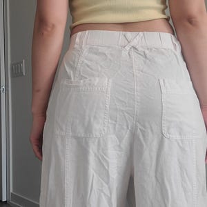 May include: Off-white, wide-leg pants with two large pockets on the back. The pants have a drawstring waist and are made of a lightweight fabric. The pants are part of a casual clothing collection.