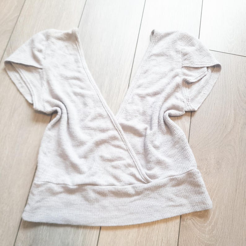 May include: A light gray wrap top with short sleeves. The top has a V-neck design and a ribbed texture. The fabric appears soft and comfortable, suitable for casual wear. The top is laid flat on a wooden surface.