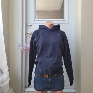 May include: A navy blue hooded sweatshirt with a kangaroo pocket and a small logo on the chest. The sweatshirt is paired with a denim mini skirt and a brown belt. The outfit is displayed on a mannequin.