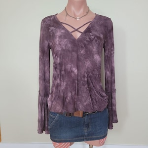 May include: A purple tie-dye style top with long sleeves and a criss-cross neckline. The top has flared sleeves and is paired with a denim mini-skirt and a brown belt. A silver necklace with a star pendant is worn.