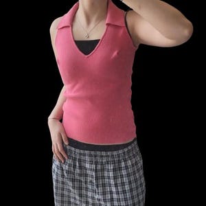 May include: A sleeveless, pink knitted top with a collared neckline and a V-neck. The top is worn over a black undershirt. The person is also wearing plaid pyjama bottoms.