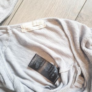 May include: Close-up of a light gray, ribbed knit top. A black tag with white text is attached to the inside of the garment. A small, off-white tag is sewn into the neckline.