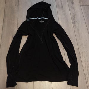 May include: Black hooded long-sleeve shirt. The shirt has a V-neck design and a hood. The hood has a light blue and white trim. The shirt is made of a soft, lightweight material. The shirt has a small logo on the chest.