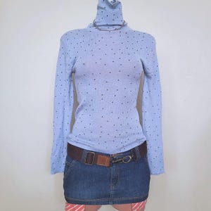 Puede incluir: Un suéter de cuello alto azul claro de manga larga con un patrón de pequeños puntos oscuros, combinado con un cinturón de tela marrón oscuro con hebilla metálica y una minifalda de mezclilla azul oscuro con costuras y dobladillo a rayas naranja.