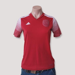 May include: A red short-sleeved jersey with a V-neckline, featuring white Adidas branding on the right chest and a tonal crest on the left. The sleeves have a white and red honeycomb pattern, and three white stripes run along the shoulders.