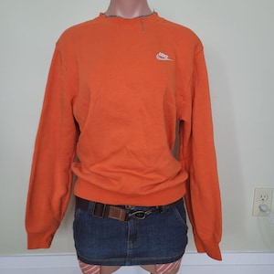 May include: Vibrant orange crew neck sweatshirt featuring a small white Nike logo on the upper left chest. The sweatshirt is styled with a denim mini skirt and a brown belt. The garment is displayed on a mannequin.