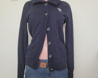Op de afbeelding: Een donkerblauw vest met lange mouwen, een geribbelde kraag, een knoopsluiting aan de voorzijde met vijf donkere knopen en een klein wit geborduurd elandlogo op de linkerborst. Het vest wordt gedragen over een blauwe spijkerrok.