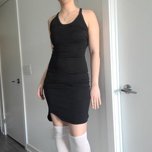 Può includere: Un abito nero senza maniche, aderente, con scollo tondo e orlo al ginocchio. L'abito ha una silhouette aderente e un leggero orlo asimmetrico. Il tessuto sembra essere un materiale morbido ed elastico. Adatto per occasioni casual o semi-formali.