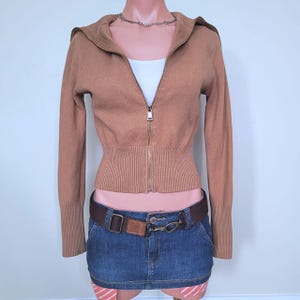 May include: A tan long-sleeve zip-up jumper with a ribbed hem and collar, worn over a white top. The jumper is paired with a dark blue denim mini skirt featuring a brown belt with a metal buckle and a silver-toned chain necklace around the neck.