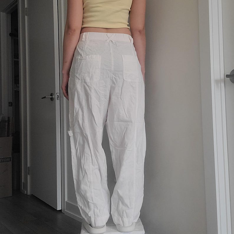 May include: White cargo pants with multiple pockets and a relaxed fit. The pants have a high waist and a slightly gathered ankle. The model is wearing a yellow crop top. The pants are made of a lightweight fabric.
