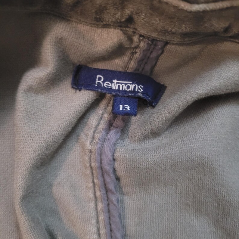 Op de afbeelding: Close-up van een kledinglabel van Reitmans. Het marineblauwe label heeft witte tekst en het nummer 13. Het kledingstuk heeft een gedempte groene kleur.