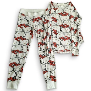 Peut inclure: Un ensemble de pyjama deux pièces composé d'un haut à manches longues et d'un pantalon assorti. Les deux pièces sont blanches et présentent un motif répétitif sur toute la surface du personnage Hello Kitty, avec un visage blanc, un nez jaune et un nœud rouge.
