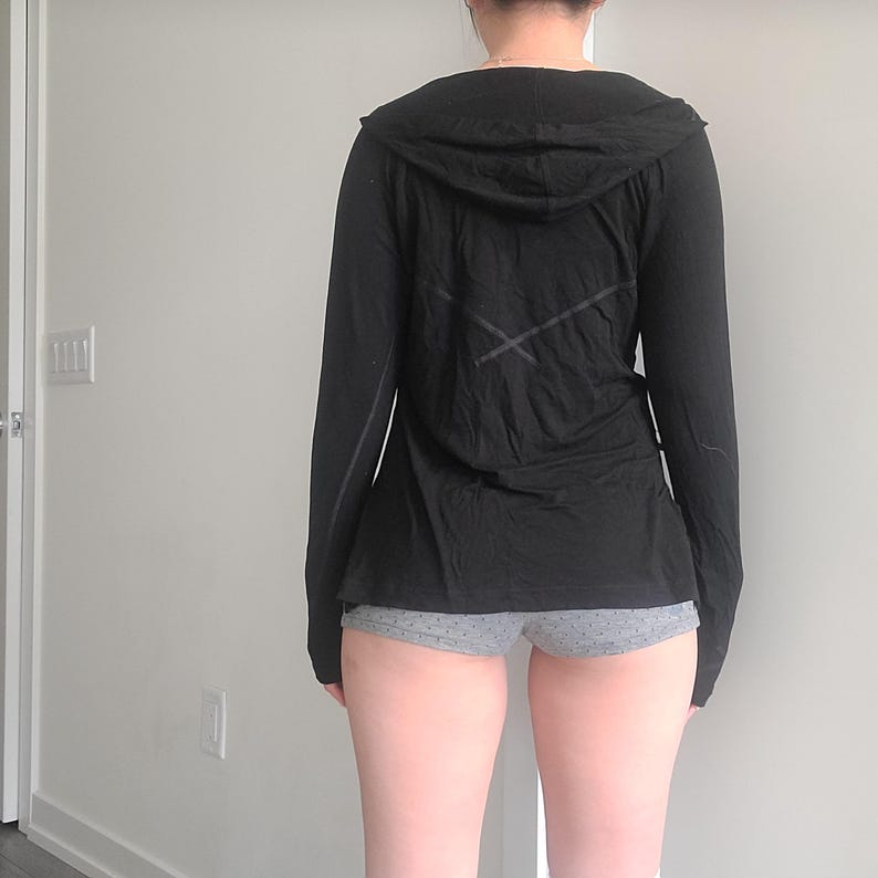 May include: Black long-sleeve hooded shirt with a cross design on the back. The shirt has a hood and a long hemline. The model is wearing gray shorts with white polka dots. The shirt is made of a soft, lightweight material.