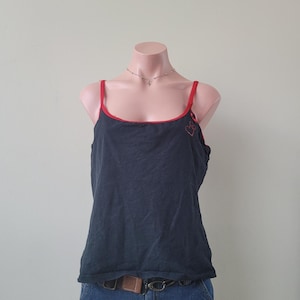 Puede incluir: Una camiseta de tirantes negra con finos tirantes rojos y un borde rojo a lo largo del escote redondo. La prenda incluye un pequeño detalle gráfico en forma de corazón rojo en la parte superior izquierda del pecho, bajo el tirante.