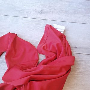 以下が含まれることがあります： 鮮やかな赤色の水着トップ、おそらくビキニスタイルが表示されています。生地は滑らかで、伸縮性のある素材のようです。トップは折りたたまれ、ネックラインとストラップが見えています。背景は明るい色の木製の表面です。