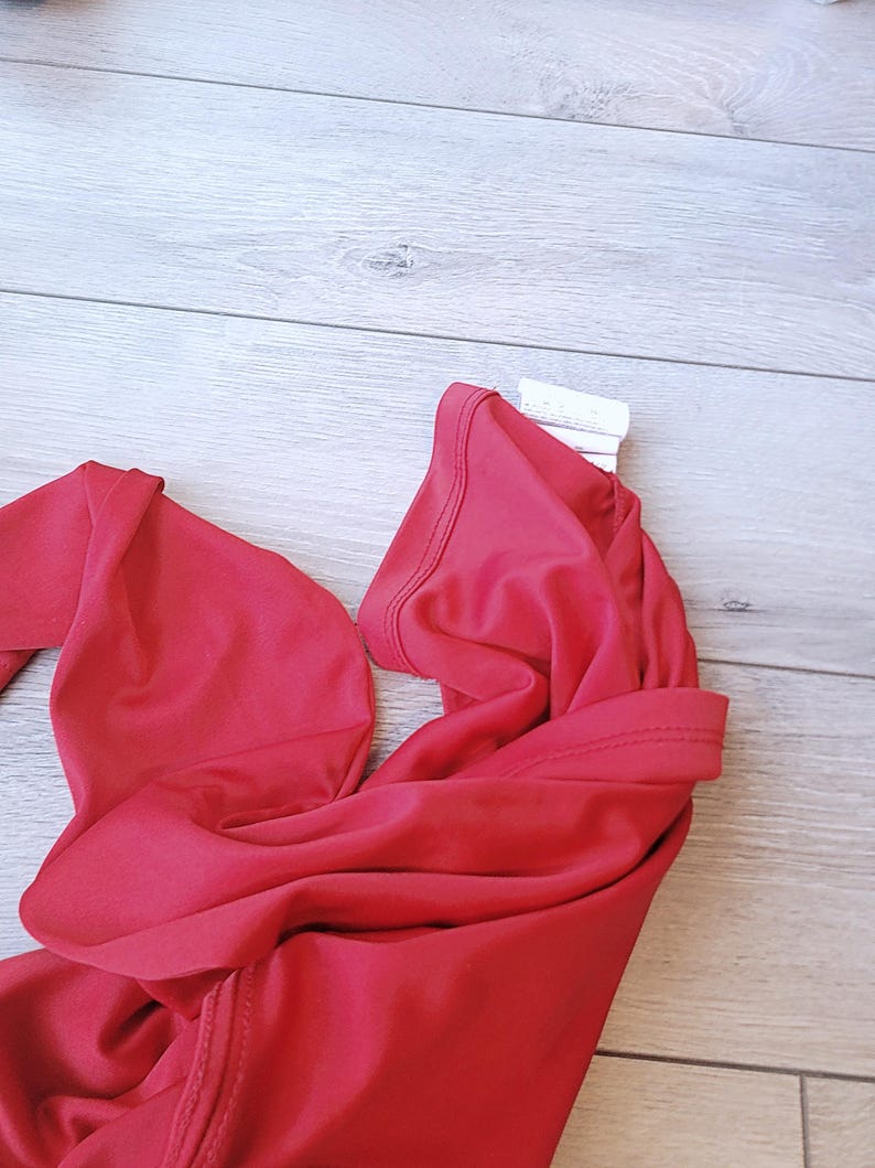 以下が含まれることがあります： 鮮やかな赤色の水着トップ、おそらくビキニスタイルが表示されています。生地は滑らかで、伸縮性のある素材のようです。トップは折りたたまれ、ネックラインとストラップが見えています。背景は明るい色の木製の表面です。