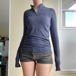 Peut inclure: Un haut de sport bleu marine à manches longues avec un col montant zippé. Le haut a une coupe ajustée et des trous pour les pouces aux poignets. Le modèle porte également un short gris foncé.