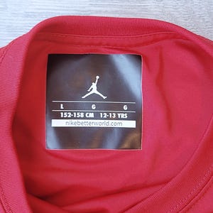 以下が含まれることがあります： 赤いスポーツシャツの襟とタグのクローズアップ。タグにはJumpmanのロゴ、サイズL、152〜158cm、12〜13歳の測定値が表示されています。ウェブサイトのアドレスはnikebetterworld.comです。
