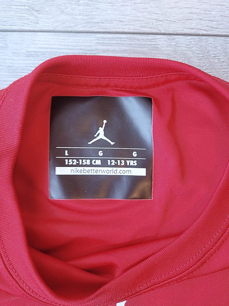 以下が含まれることがあります： 赤いスポーツシャツの襟とタグのクローズアップ。タグにはJumpmanのロゴ、サイズL、152〜158cm、12〜13歳の測定値が表示されています。ウェブサイトのアドレスはnikebetterworld.comです。