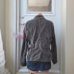 Op de afbeelding: Een grijze corduroy jas met een knoopsluiting en een ceintuurdetail in de taille. De jas wordt getoond op een mannequin met een spijkerbroekrok. De jas heeft een klassiek, getailleerd ontwerp.