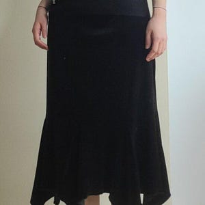 Puede incluir: Una falda midi negra con un dobladillo único y un top negro a juego. La falda tiene un diseño acampanado y asimétrico. La modelo lleva calcetines amarillos. El atuendo es sencillo y elegante.