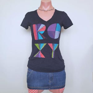 May include: A black short-sleeved V-neck t-shirt featuring the word ROXY printed in large, colourful, geometric patterns. The letters are filled with various textures and shades of red, blue, purple, and green. The shirt is paired with a blue denim skirt.