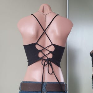Op de afbeelding: Een zwarte croptop met een vetersluiting aan de achterkant, waarbij dunne zwarte koorden kruislings lopen en aan de onderkant worden vastgemaakt. Het kledingstuk wordt getoond op een paspop en toont de open rug en het verstelbare sluitsysteem.