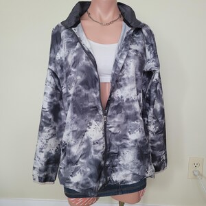 Peut inclure: Veste à imprimé tie-dye noir et blanc avec fermeture éclair complète. La veste a un col et des manches longues. Un haut blanc est visible en dessous. La veste est présentée sur un mannequin.