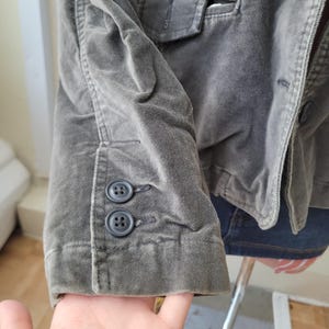 Op de afbeelding: Close-up van een grijsgroene corduroy blazer. De mouwboord heeft twee donkergrijze knopen. De stof heeft een zacht, getextureerd uiterlijk. De blazer maakt deel uit van een kledingcollectie.