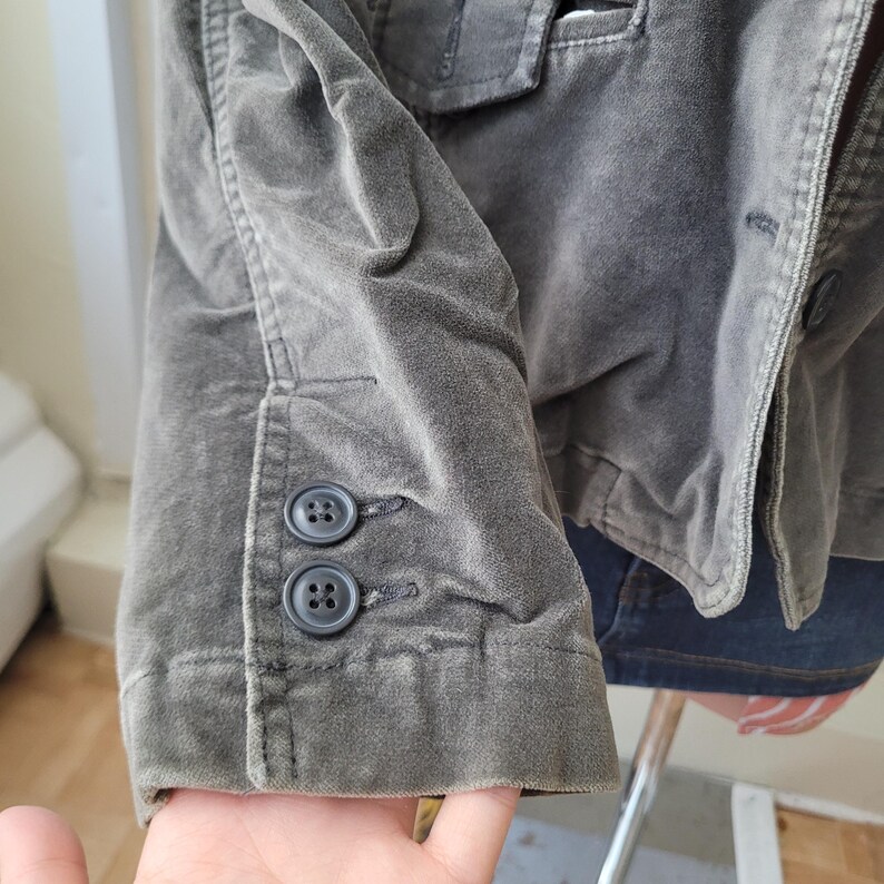 Op de afbeelding: Close-up van een grijsgroene corduroy blazer. De mouwboord heeft twee donkergrijze knopen. De stof heeft een zacht, getextureerd uiterlijk. De blazer maakt deel uit van een kledingcollectie.