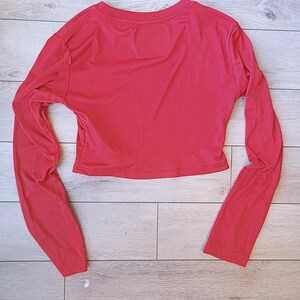 以下が含まれることがあります： 鮮やかな赤色のクロップド丈長袖トップス。クルーネックで、体にフィットするシルエット。袖は長く、手首まであります。生地は滑らかで軽く、カジュアルな着こなしに適しています。