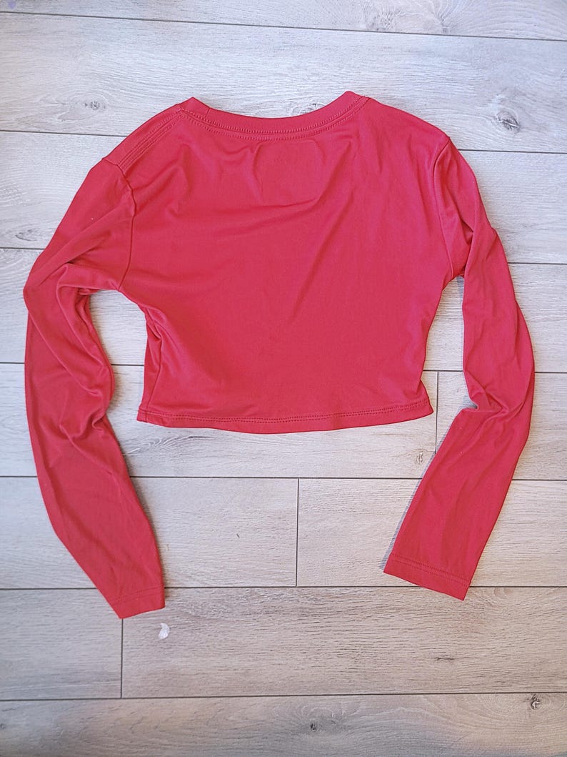 以下が含まれることがあります： 鮮やかな赤色のクロップド丈長袖トップス。クルーネックで、体にフィットするシルエット。袖は長く、手首まであります。生地は滑らかで軽く、カジュアルな着こなしに適しています。