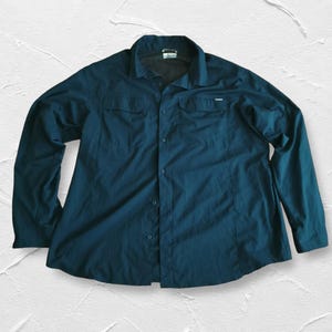May include: A dark teal long-sleeve button-down shirt featuring a classic collar, two chest pockets with flaps, and a central button placket. The garment is constructed from a smooth, lightweight fabric and includes a small brand tag on the chest.