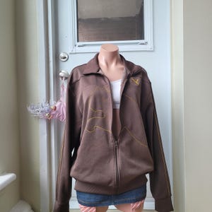 May include: A brown zip-up jacket with yellow stitching details and a small yellow logo on the chest. The jacket is displayed on a mannequin. The jacket has a collar and long sleeves.
