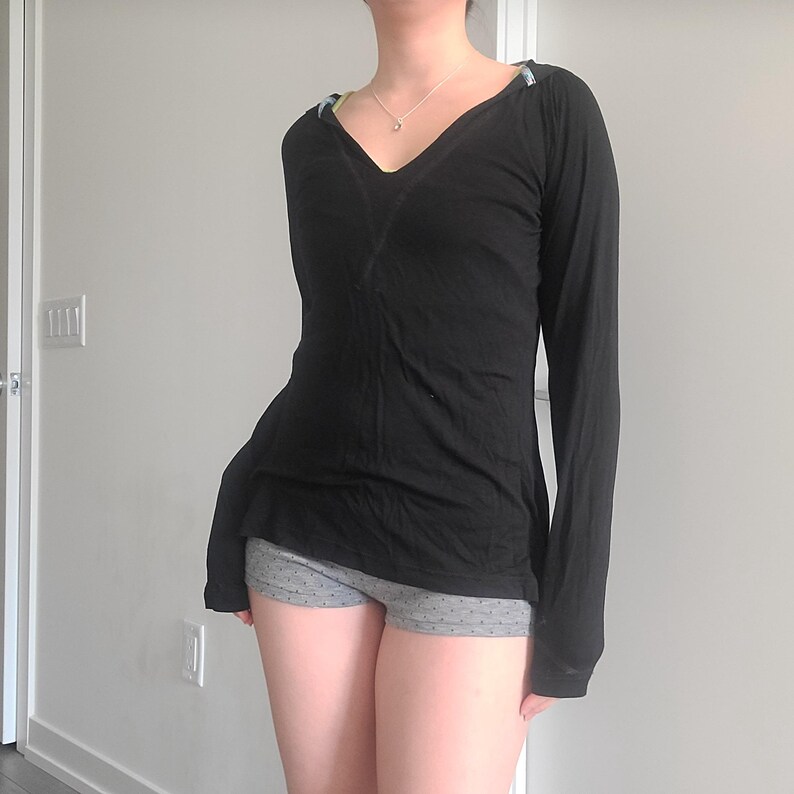 May include: A black long-sleeve shirt with a V-neck design. The shirt has a fitted silhouette and is paired with gray shorts with a small dot pattern. The shirt has a decorative detail on the shoulders.